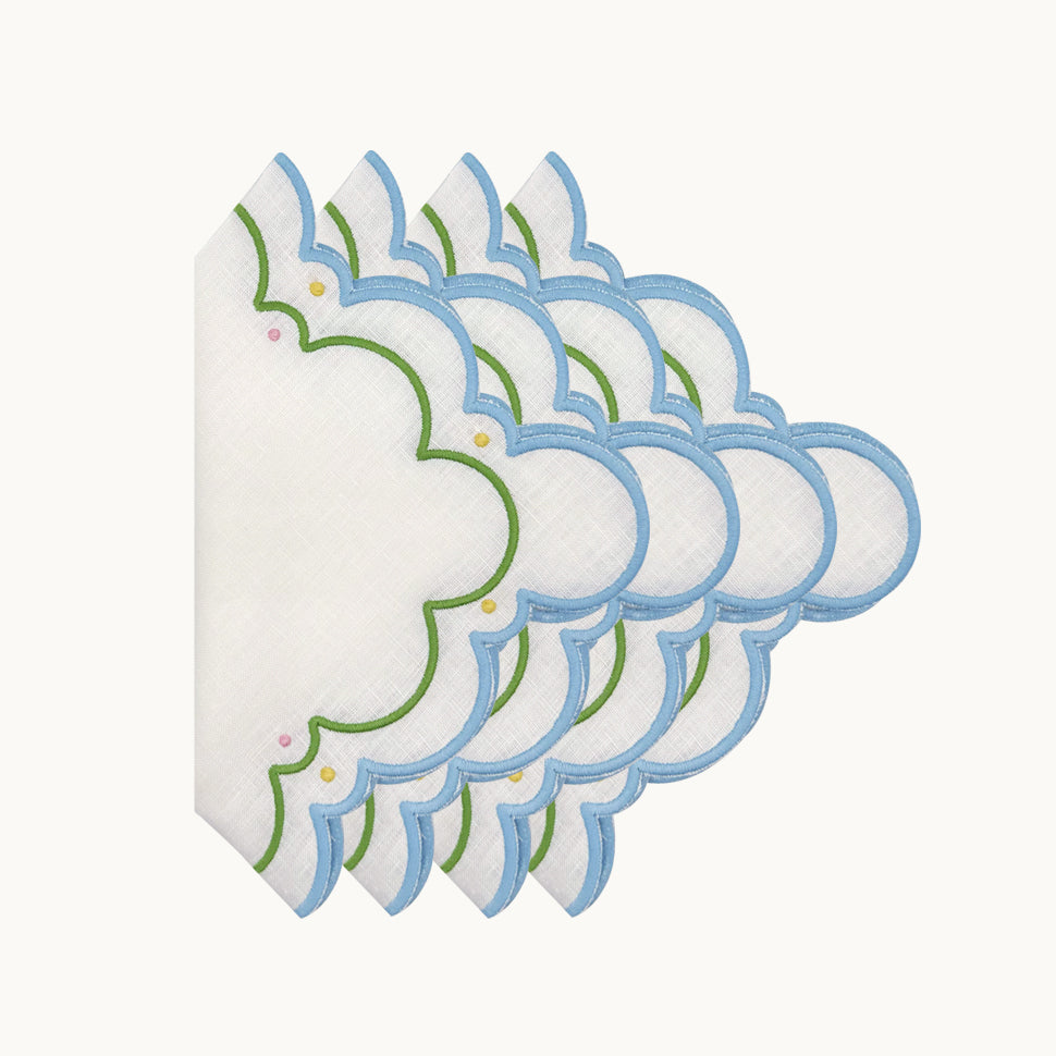 A stack of Sugus Table Linen Collection napkins, featuring white linen with scalloped edges embroidered in blue, green, and small yellow and pink dots.