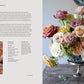 An exquisite arrangement from Penguin Random House’s “A Sweet Floral Life” showcases assorted roses in pink, peach, yellow, and cream hues displayed in a dark metal vase on a table, beautifully capturing the artistry found within expert floral designs, with softly blurred plates visible in the background.