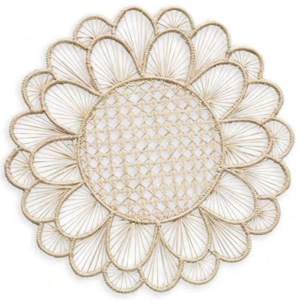 The Symi Rattan Placemat Collection by Themis Z features round, handcrafted placemats with a flower-like pattern, tightly woven center, and petal-shaped edges made from natural-colored fibers by Colombian artisans.