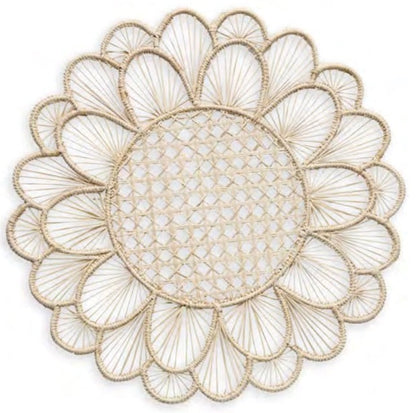 The Symi Rattan Placemat Collection by Themis Z features round, handcrafted placemats with a flower-like pattern, tightly woven center, and petal-shaped edges made from natural-colored fibers by Colombian artisans.