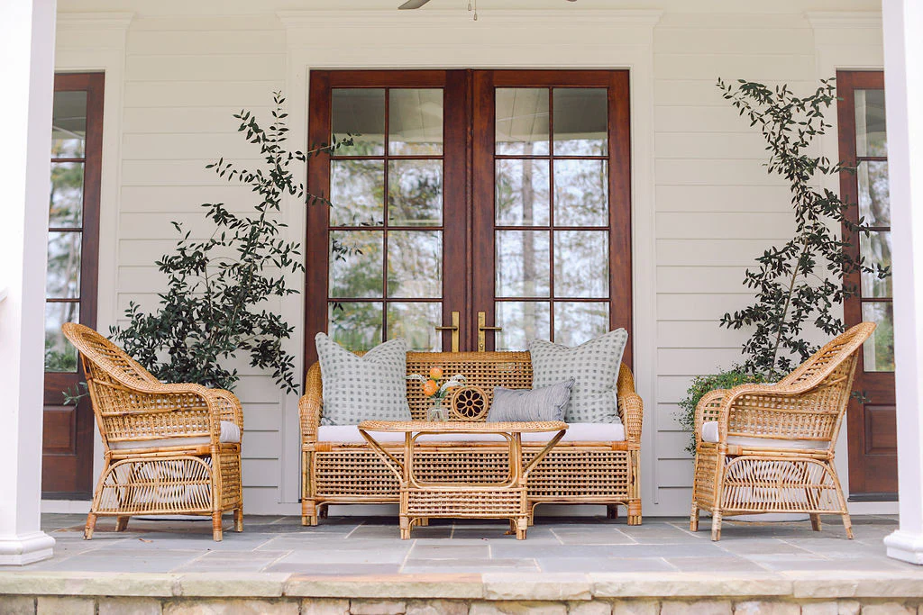 A porch featuring the Varnish Collection's Julianna Sofa, two artisan wicker chairs, and a glass-top coffee table, set by double wooden doors and flanked with two potted plants crafted from natural materials.