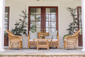 A porch featuring the Varnish Collection's Julianna Sofa, two artisan wicker chairs, and a glass-top coffee table, set by double wooden doors and flanked with two potted plants crafted from natural materials.