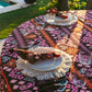 A round table set for two features Figue’s Dancing Ikat Ganache Collection, with artisan-crafted plates, glasses, cutlery, and pink-orange ikat table linens, outdoors by a garden and pool.