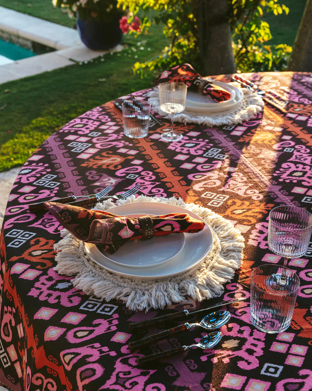 A round table set for two features Figue’s Dancing Ikat Ganache Collection, with artisan-crafted plates, glasses, cutlery, and pink-orange ikat table linens, outdoors by a garden and pool.
