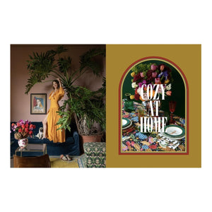 A woman in a yellow La DoubleJ dress stands in a plant-filled living room next to Table Energy: Entertaining with Italian Spirit by Abrams on the decorated dining table.