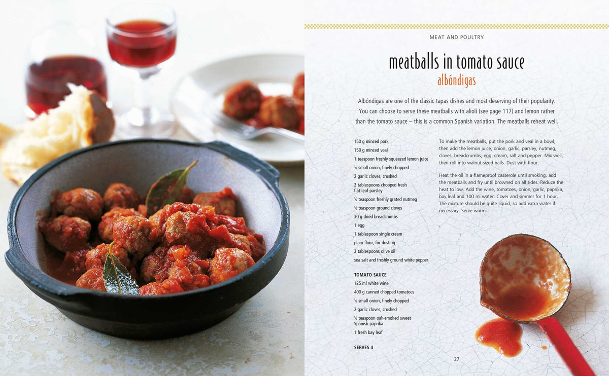 A bowl of meatballs in tomato sauce with bay leaves—a classic among Spanish appetizers—takes center stage, while a glass of red wine sits in the background and an open page from *Tapas* by Simon & Schuster showcases one of the beloved recipes for meatballs in tomato sauce.