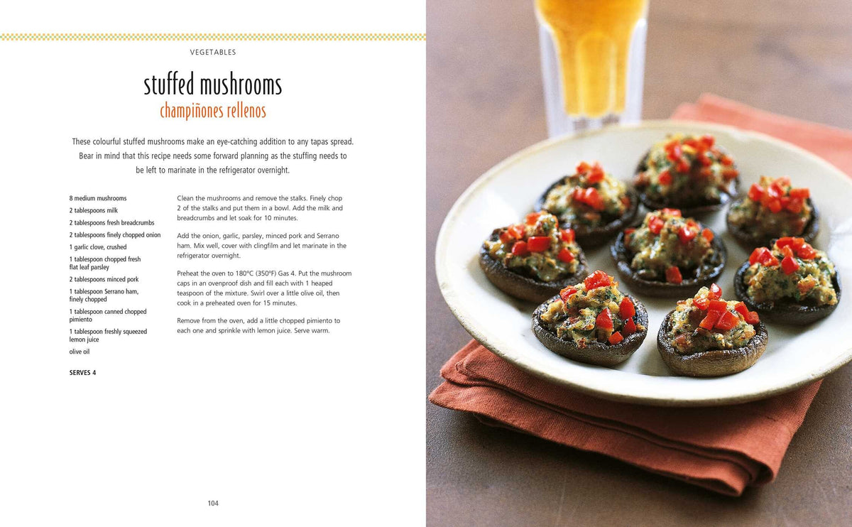 Six stuffed mushroom caps, topped with chopped red peppers, are artfully presented on a dark plate beside a glass of light-colored beverage—the perfect Spanish appetizer from the “Tapas” collection by Simon & Schuster, as shown on the left.