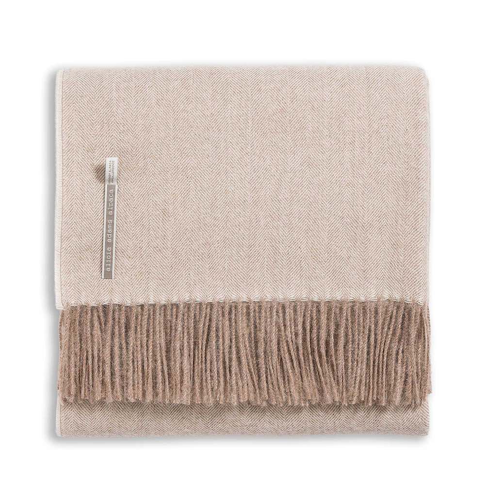 Alicia Adams Alpaca Classic Herringbone Throw Collection: folded beige alpaca blanket with brown fringe and a small label near the edge, fair-trade made in Peru.