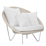 The Janus et Cie Lounge Chair in Schooner Snow features a round beige wicker frame, metal legs, plush cushion, and three large white pillows—ideal for adding midcentury modern elegance to your luxury space.