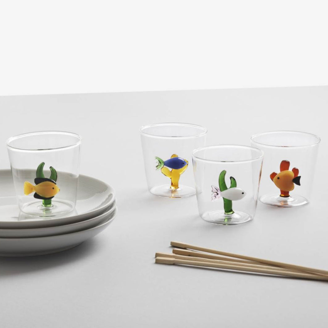 Four glass tumblers from Ichendorf Milano's Marine Garden Drinkware Collection, each featuring handmade glass fish, are arranged near stacked white plates and a pair of chopsticks on a white surface.