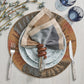 A place setting with a fork, knife, patterned plates, two blue glasses, a multicolored napkin in the hand-carved Kim Seybert Tendril Napkin Ring, and decorative branches on a light wood table.