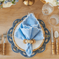 A wooden table set for formal dining features Kim Seybert’s Arches Napkin Set with embroidered edges, bamboo-handled cutlery, a blue scalloped placemat, a white plate in a napkin ring, and two crystal glasses.