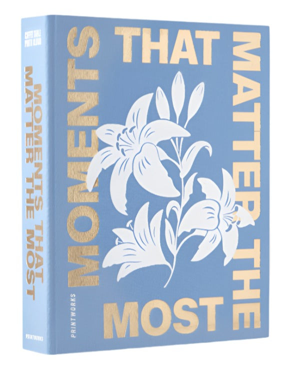The PrintWorks "Moments That Matter The Most" Photo Album features a blue cotton cover with gold text and a white lily illustration on the front.