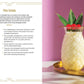 A refreshing Mocktail Club piña colada is served in a pineapple-shaped glass, garnished with leaves and cherries, and placed on a gold tray alongside a Simon & Schuster recipe book featuring delicious alcohol-free cocktails and instructions—all set against a vibrant pink and purple background.