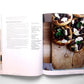 An open copy of “Ottolenghi, The Cookbook” by Penguin Random House displays a Mediterranean recipe on the left page, while the right page features a photo of small round pizzas with toppings arranged on a wooden board.