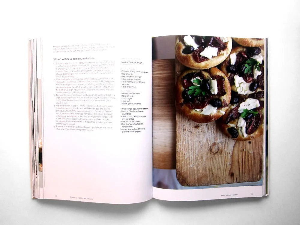 An open copy of “Ottolenghi, The Cookbook” by Penguin Random House displays a Mediterranean recipe on the left page, while the right page features a photo of small round pizzas with toppings arranged on a wooden board.