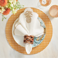 A table setting featuring a gold placemat, white plate, leaf side plate, Kim Seybert Pina Punch Napkin Set with gold embroidery, a wooden napkin ring, and elegant glassware.
