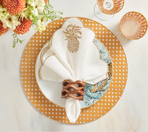 A table setting featuring a gold placemat, white plate, leaf side plate, Kim Seybert Pina Punch Napkin Set with gold embroidery, a wooden napkin ring, and elegant glassware.