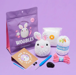 The Woobles Jojo the Bunny Beginner Crochet Kit comes with loopable yarn, stuffing, a crochet hook, pattern booklet, buttons, and a finished Jojo the Bunny—each item neatly displayed on a purple surface.
