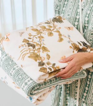 A person holds two folded Vivian Magnolia Floral Scallop Towels from Weezie Towels, featuring a green geometric and floral design, while wearing a green and white patterned garment.
