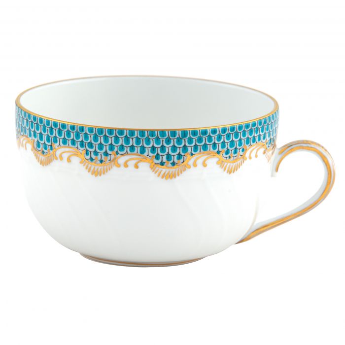 A white porcelain teacup from Herend’s Fish Scale Turquoise Collection features a gold rim and handle with a blue and gold scalloped pattern, echoing the hand-painted elegance of Herend dinnerware.
