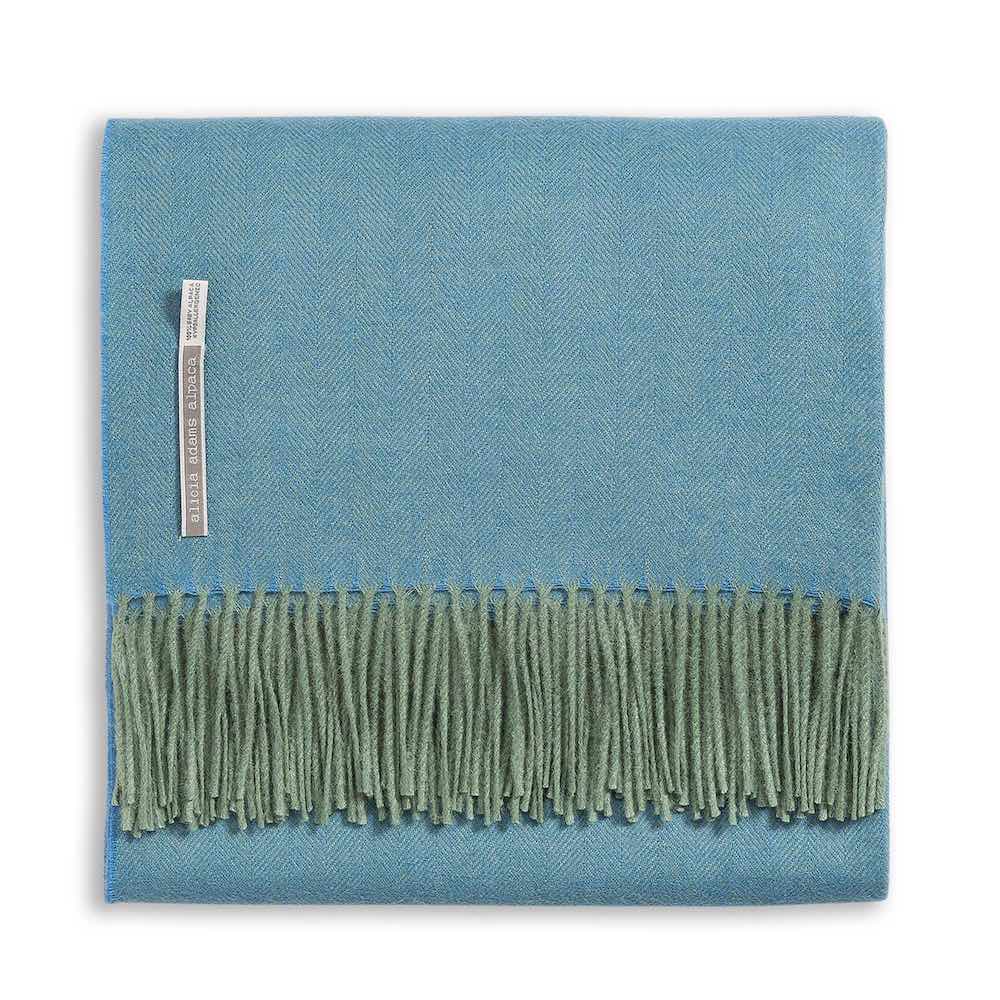 The Alicia Adams Alpaca Classic Herringbone Throw Collection features a folded blue herringbone-patterned baby alpaca blanket with a green fringe and fabric label, fair-trade crafted in Peru.