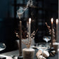 A dark dining table adorned with elegantly lit Baobab Collection Candle Holder Twins, glistening wine glasses, sophisticated table settings, featuring exquisite hand-blown glass vases filled with lush foliage.