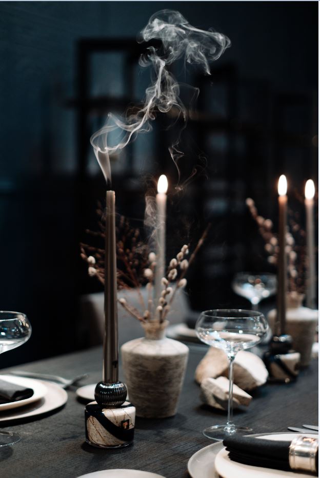 A dark dining table adorned with elegantly lit Baobab Collection Candle Holder Twins, glistening wine glasses, sophisticated table settings, featuring exquisite hand-blown glass vases filled with lush foliage.