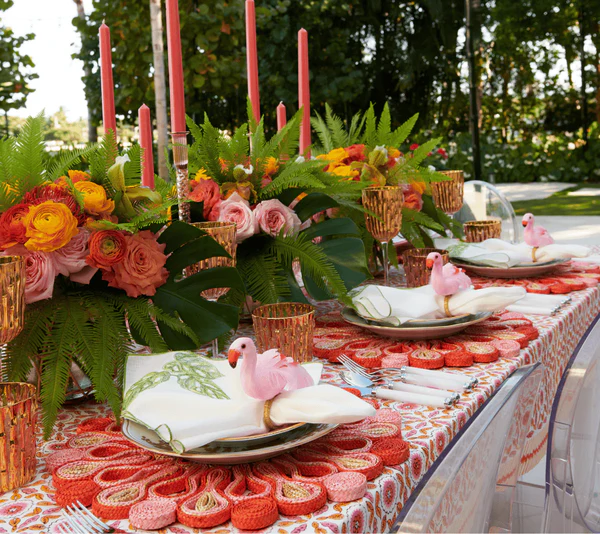 A decorated outdoor table features floral centerpieces, pink candles, gold-rimmed glasses, and plates with napkins topped by the Kim Seybert Aruba Napkin Ring in Blush.