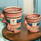 Two Baobab Collection Fety Mandihy scented candles in pink, blue, and beige woven holders sit on a wooden table. Each has multiple wicks and a rectangular label against a light blue background, enhancing their charm.
