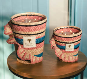 Two Baobab Collection Fety Mandihy scented candles in pink, blue, and beige woven holders sit on a wooden table. Each has multiple wicks and a rectangular label against a light blue background, enhancing their charm.