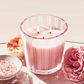 A Nest Himalayan Salt & Rosewater Candle with pink stripes and two lit wicks sits on glass, its amber and rosewater scent in the air. Pink bath salts and flowers nearby complete the elegant scene from the collection.