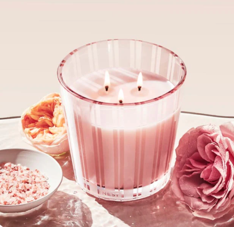 A Nest Himalayan Salt & Rosewater Candle with pink stripes and two lit wicks sits on glass, its amber and rosewater scent in the air. Pink bath salts and flowers nearby complete the elegant scene from the collection.