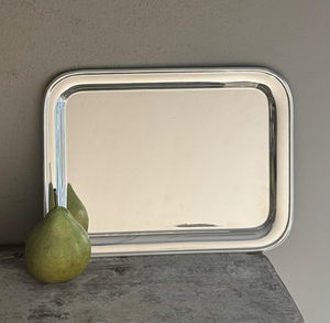 A Vintage Oblong Tray by Hôtel Silver stands upright against a wall on a wooden surface, with a green pear placed in front of it.