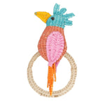 The Cockatoo Napkin Ring by Mercedes Salazar features a handmade woven toy bird with a blue head, yellow beak, pink and orange body, and long tail, all crafted from raffia fiber and attached to a beige woven ring in Colombia.