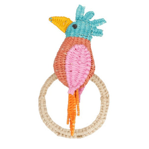 The Cockatoo Napkin Ring by Mercedes Salazar features a handmade woven toy bird with a blue head, yellow beak, pink and orange body, and long tail, all crafted from raffia fiber and attached to a beige woven ring in Colombia.