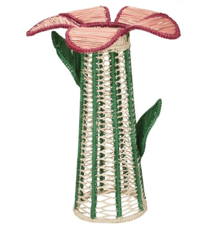 Handcrafted from natural raffia fiber in the signature style of Mercedes Salazar, the Lily Floral Spirit Candle Holder & Flower Vase features a woven wicker sculpture resembling a tall flower with pink petals and green leaves on a cream and green cylindrical stem.