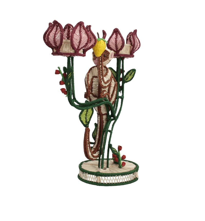 The Jungle Whisper Mico Candle Holder by Mercedes Salazar features a handwoven raffia elephant figurine standing amid three pink flowers with green stems, all crafted from colorful thread on a circular base—an enchanting example of artisanal decor.