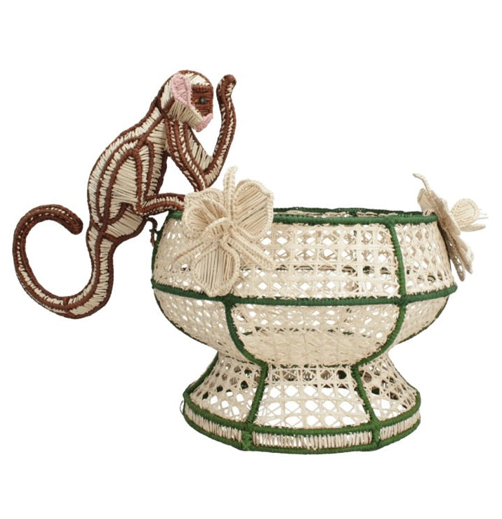 Handcrafted from raffia, the Forest Muse Mono Bowl by Mercedes Salazar features a woven design with a monkey figure climbing on the rim and two butterfly ornaments attached to the side. Inspired by vibrant designs from Bogotá, Colombia, this decorative basket is accented in green.