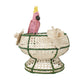 The Tropical Harmony Cockatoo Bowl by Mercedes Salazar is a woven decorative bowl handcrafted from natural raffia fiber, featuring green accents, two cream flowers, and a pink parrot with a yellow crest perched on the rim—an exquisite piece inspired by the artisanal traditions of Bogotá, Colombia.