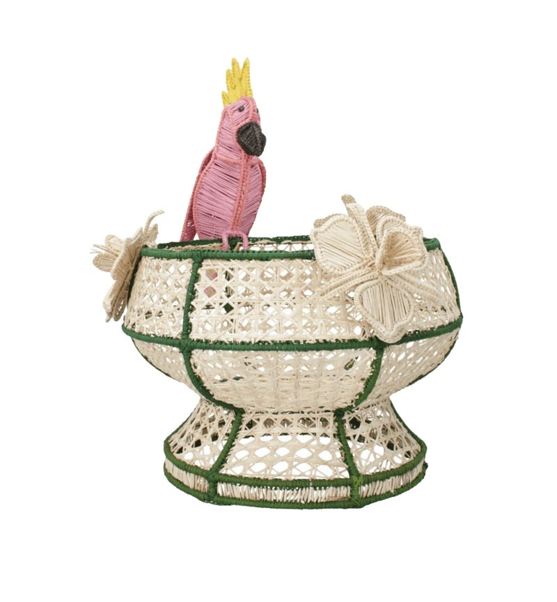 The Tropical Harmony Cockatoo Bowl by Mercedes Salazar is a woven decorative bowl handcrafted from natural raffia fiber, featuring green accents, two cream flowers, and a pink parrot with a yellow crest perched on the rim—an exquisite piece inspired by the artisanal traditions of Bogotá, Colombia.