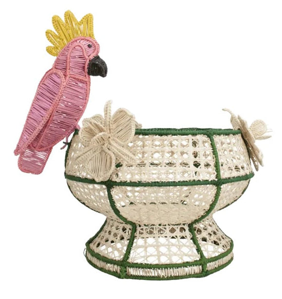 The Tropical Harmony Cockatoo Bowl by Mercedes Salazar is a handcrafted woven basket made from natural raffia fiber in Bogotá, Colombia, featuring green trim, a pink and yellow parrot motif, and two decorative flowers attached to the side.