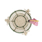 The Tropical Harmony Cockatoo Bowl by Mercedes Salazar is a woven round basket handcrafted from natural raffia fiber in Bogotá, Colombia, featuring a green rim, four decorative bows, and a pink and yellow butterfly perched on the edge, as seen from above.
