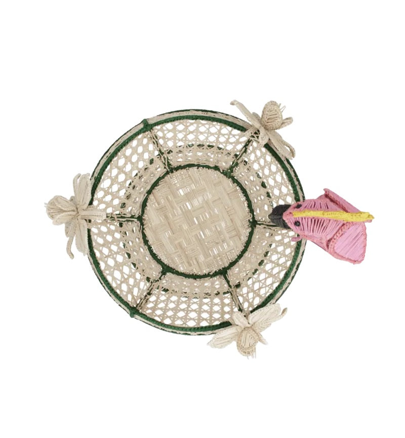 The Tropical Harmony Cockatoo Bowl by Mercedes Salazar is a woven round basket handcrafted from natural raffia fiber in Bogotá, Colombia, featuring a green rim, four decorative bows, and a pink and yellow butterfly perched on the edge, as seen from above.
