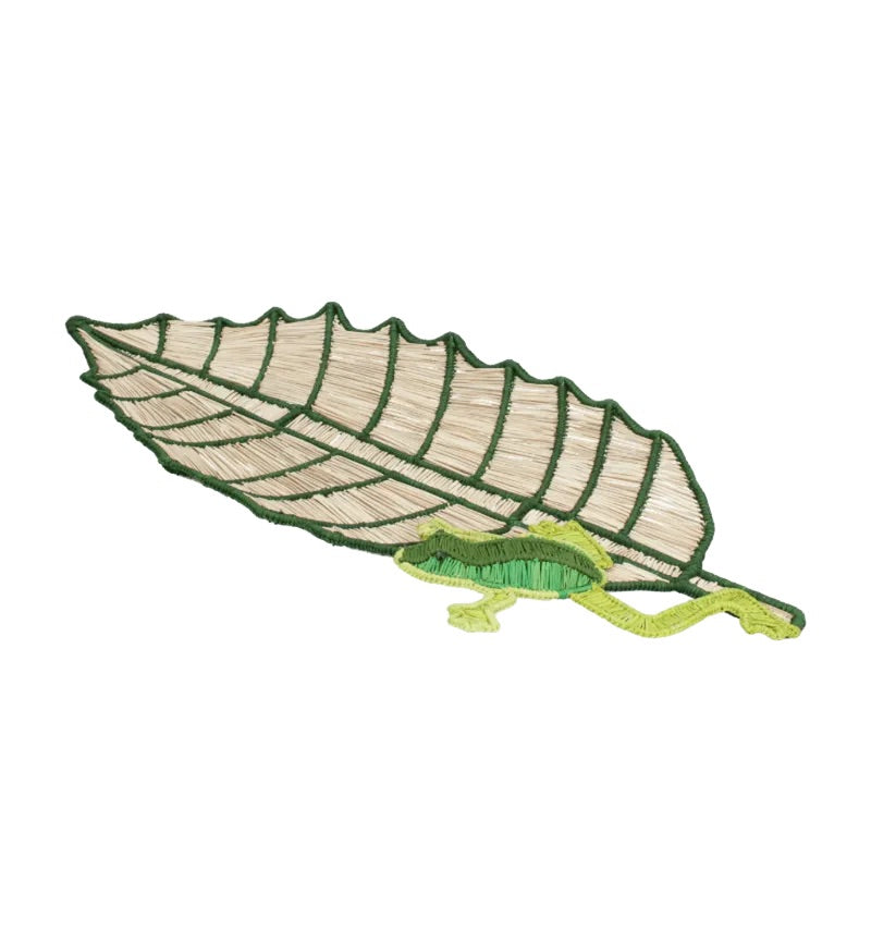 The Frog Leaf Amazonas Tray by Mercedes Salazar features an embroidered patch of a green leaf with a small green lizard beneath it, echoing the natural motifs found in handwoven raffia fiber trays, set against a plain white background.