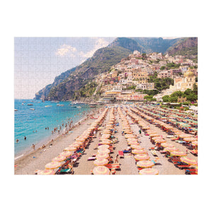 The Italy Two-Sided Puzzle by Chronicle Books features a lively beach scene with colorful umbrellas, sunbathers, hillside buildings, and mountains, capturing the charm of an Italian coast.