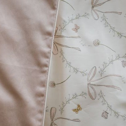 A close-up of Gooselings' Bows & Butterfly Kisses Toddler Duvet shows solid light pink fabric paired with white featuring a delicate bow, floral, and butterfly pattern—ideal for a lightweight toddler duvet.