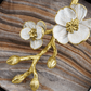 The Michael Aram Orchid Cocktail Napkin Holder features a gold and white metal floral design with two blossoms and three buds, resting on brown, beige, and gray striped fabric—a touch of subtle luxury and elegance.