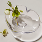 The Simon Pearce Romance Heart Dish features a unique heart-shaped design holding a green and white flower with leaves and buds, plus a matching flower beside it—ideal as romantic table decor or an elegant jewelry holder.