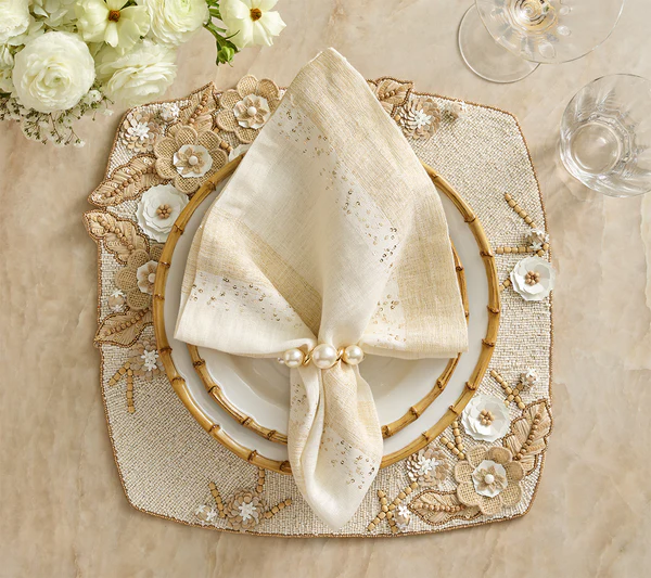 A formal spring table features a Kim Seybert Bramble Placemat in Ivory/Natural, white plates, a gold-rimmed napkin ring with a folded napkin, and a bouquet of white flowers alongside elegant glassware.